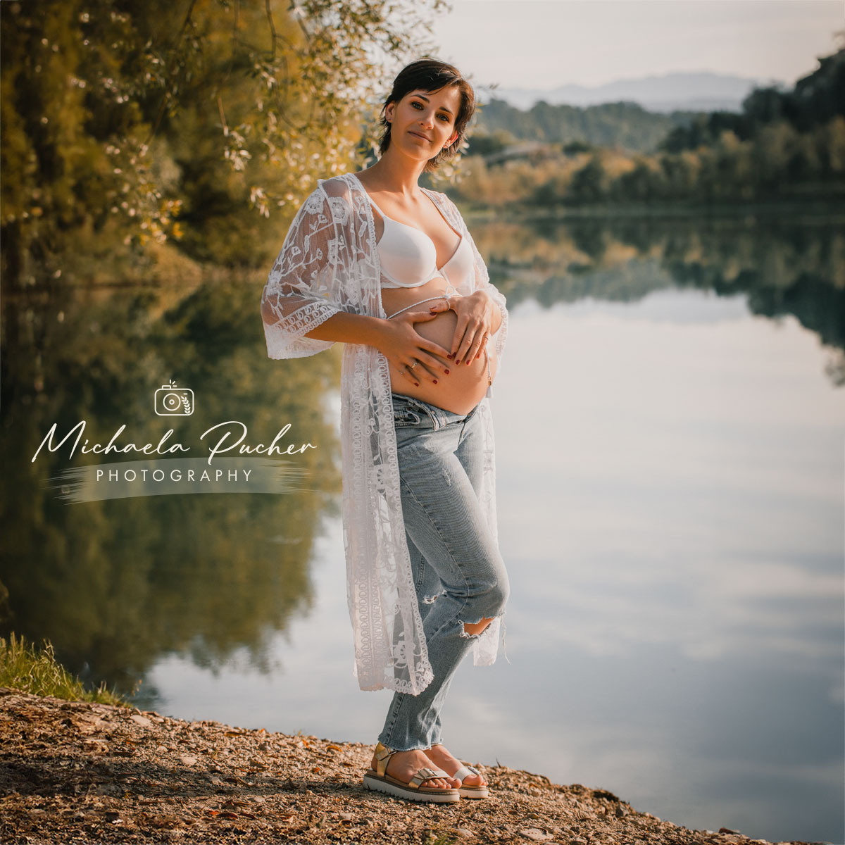 Schwangere Frau steht entspannt am Seeufer, hält sanft ihren Babybauch und lächelt in die Kamera; sie trägt Jeans, ein weißes Top und einen Spitzenkimono.